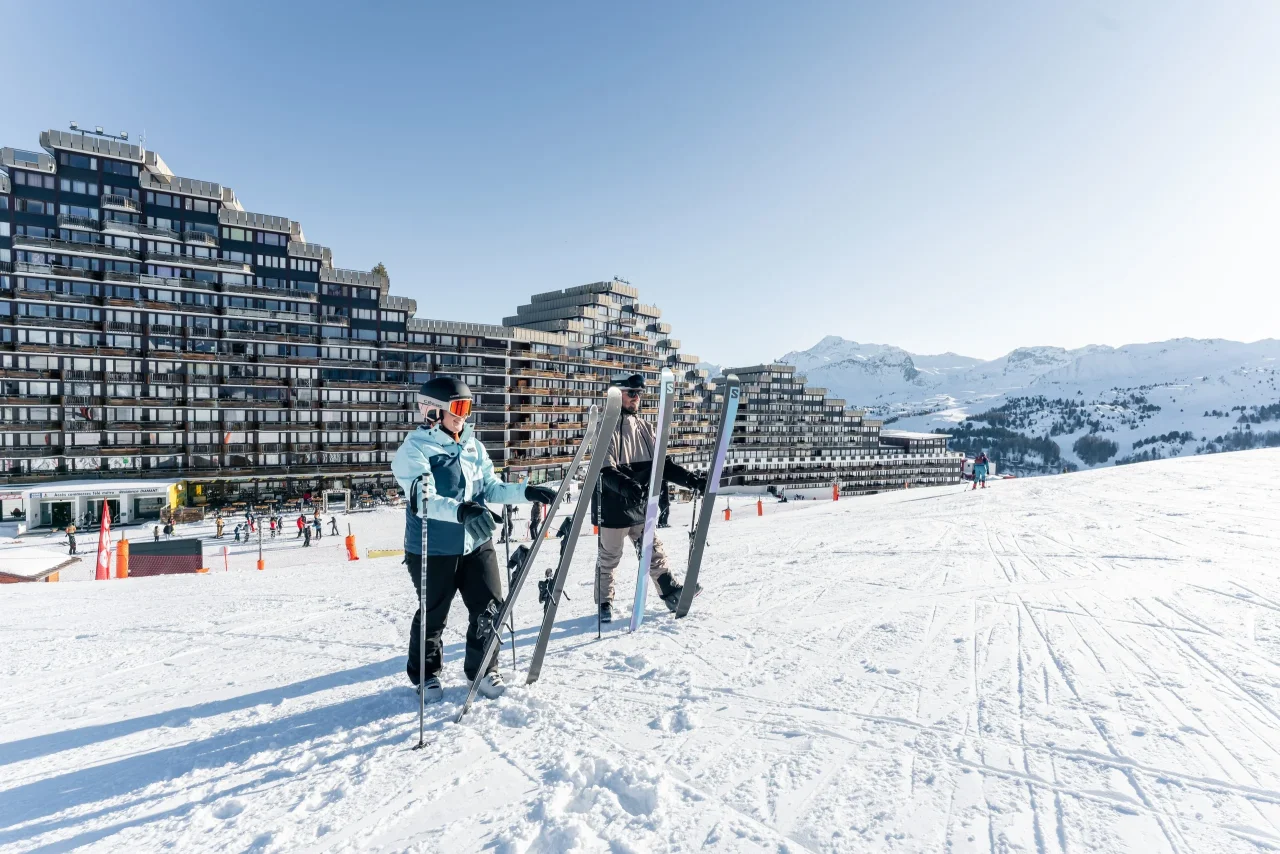 Skiers devant immeubles et montagnes enneigées, ciel bleu.